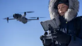 Woman in a fur-lined hood controls a flying drone in cold weather, looking at a tablet.